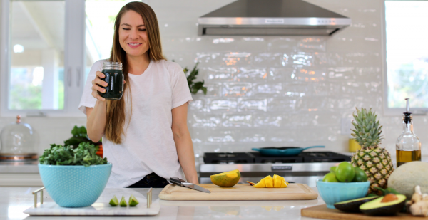 Chrissy Weir stands in her kitchen and smiles with a dark beverage in hand. A cutting board with mangoes, along with other fruits and vegetables adorn the countertop.