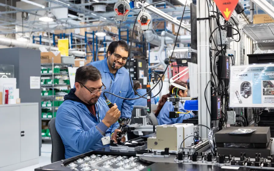 Two men in blue coats use an electronic tool on an assembly line