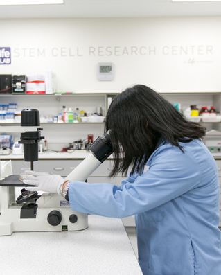 A woman in a blue lab coat peers into a microscope in a lab