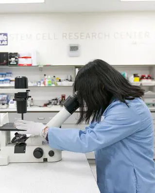 A woman in a blue lab coat peers into a microscope in a lab
