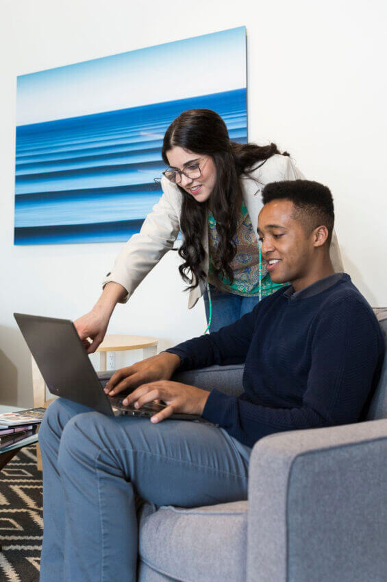 A woman leans over a seated man, pointing to something on his laptop screen